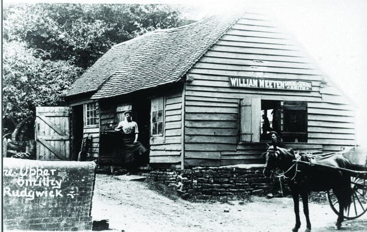 The Upper Smithy at Cox Green: a weatherboarded forge with a tiled roof, the blacksmith standing in the doorway and a horse harnessed to a cart outside. A painted sign above the door reads William Meeten.